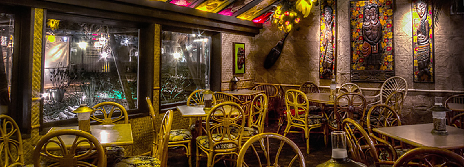 The dining room at Hala Kahiki, lit by a stained-glass tiki-pattern ceiling, with rattan chairs, carved tiki wall panels, and a view out to the patio