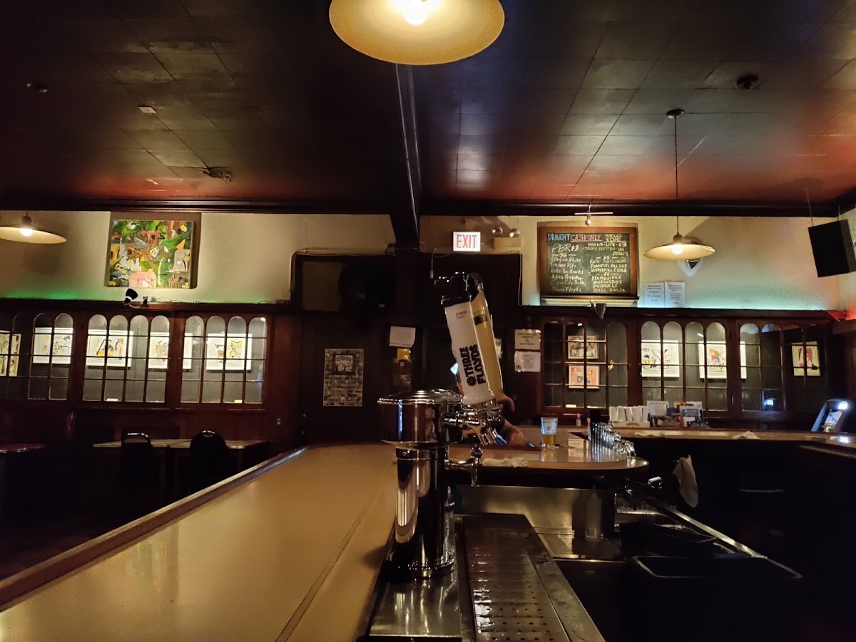 The bar at Rainbo Club in Chicago, with a zigzagging wood counter, framed local art, string lights, and a chalkboard drink menu
